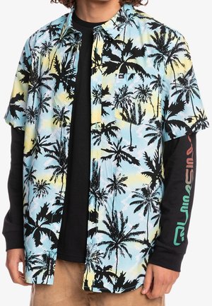 Person iført en kortærmet skjorte med blå, gul og sort palmeprint over en sort langærmet skjorte.