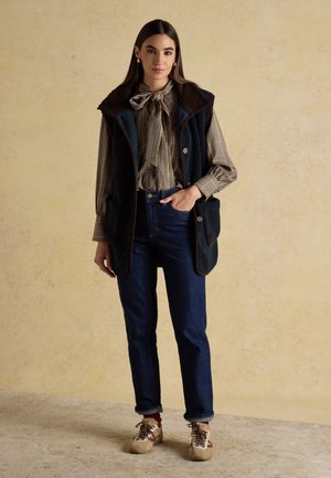 Donna in piedi che indossa una blusa a quadri con fiocco, gilet in pile nero senza maniche, jeans scuri e sneakers beige con lacci su sfondo neutro.