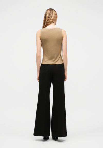 Γυναίκα με κυματιστά ξανθά μαλλιά, που φοράει αμάνικη μπεζ μπλούζα και μαύρο παντελόνι φαρδύ στο κάτω μέρος, στέκεται γυρισμένη με την πλάτη σε ένα απλό φόντο.