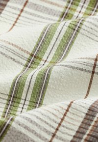 Tissu blanc à motif écossais tissé avec des rayures vertes, marron et beige, montrant la texture et de légers plis dans le matériau.