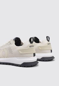 Zapatillas deportivas beige con parte superior de malla texturizada, suela negra en contraste y acentos de logo en el talón. Cuentan con una lengüeta para facilitar el uso.