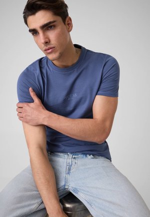 Giovane uomo con una maglietta blu Guess e jeans azzurri chiaro seduto su uno sgabello con la mano sinistra che tiene il braccio destro.