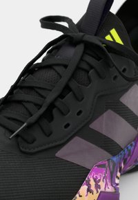 Gros plan d'une basket noire avec des lacets noirs, des rayures gris foncé, un logo jaune néon et une semelle multicolore à motif violet.