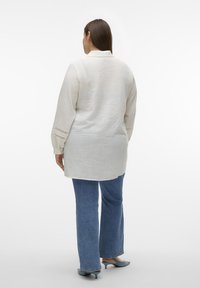 Camisa de manga larga color beige claro con tela texturizada, un cuello y costuras detalladas en los puños, combinada con unos vaqueros acampanados azules.