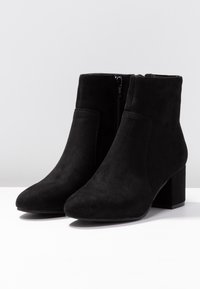Botas de tobillo de ante negro con tacones cuadrados y cremalleras laterales, mostradas en un par sobre una superficie blanca con un fondo claro y sencillo.