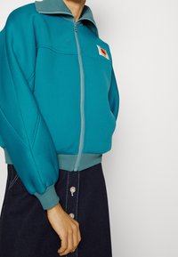 Tealfärgad zip-up jacka med texturerat tyg, ribbade ärmslut och krage. Har en patch-logo och en kontrasterande dragkedja. Kombination med jeanskjol.