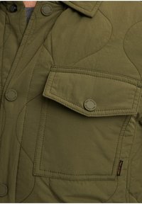 Olivgrüne Steppjacke mit einem Knopfkragen, großer Brusttasche und runden Knöpfen mit einem Markenlogo auf der Vorderseite.