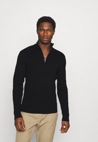 Black zip-neck sweater with ribbed texture, long sleeves, and a silver zipper. Paired with beige trousers, against a plain white background.
