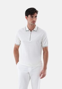 Weißes Poloshirt mit halber Reißverschluss, kontrastierendem schwarzem gestreiftem Kragen und Ärmeln, aus leichtem Stoff, getragen mit weißen Hosen.