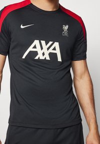 Svart sporttröja med röda axelaccenter. Har Nike-logga, stor vit "AXA"-text och Liverpool Football Club-crest. Texturerad tyg.
