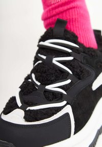 Sneaker nero con accenti bianchi, tomaia in tessuto morbido e suola spessa. Presenta lacci a contrasto e una superficie testurizzata. Calzino rosa brillante visibile.