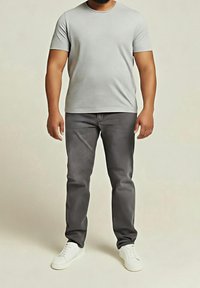 Homme debout portant un t-shirt gris clair à manches courtes, un jean gris foncé et des baskets blanches, sur un fond uni clair.