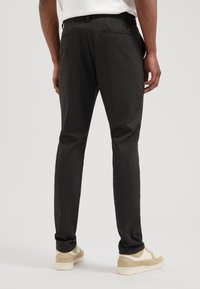 Donkergrijze broeken met een slim fit, voorzien van twee achterzakken met knoopsluitingen en een gladde textuur. Gecombineerd met lichtgekleurde schoenen.