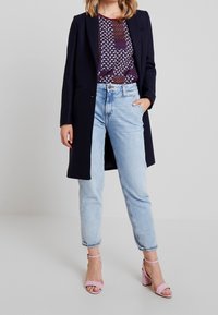 Abrigo navy, blusa estampada en tonos de púrpura y marrón, vaqueros de cintura alta en azul claro, y sandalias de tacón cuadrado rosa.