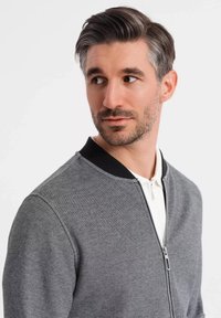 Šedá bomber bunda na zip s texturovaným povrchem, černým žebrovaným límcem a bílou košilí pod ní. Jednoduchý, moderní design, žádné viditelné kapsy.