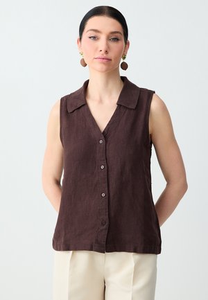 Femme aux cheveux foncés portant des boucles d'oreilles dorées et marron ainsi qu'une chemise sans manches marron foncé boutonnée, debout devant un fond blanc.