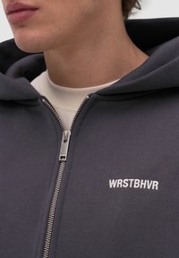 Primer plano de una persona que lleva una sudadera con capucha gris oscuro con cremallera y "WRSTBHVR" bordado en el pecho, y una camiseta blanca debajo.