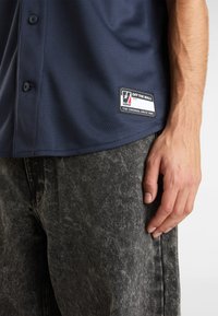 Marineblauw mesh overhemden met een label met een logo; gecombineerd met vervaagde zwarte denim broeken, die een gestructureerd oppervlak tonen.