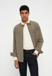 Olijfgroene shirtjas met twee grote voorkruiszakken, knoopsluiting en lange mouwen. Draag het over een lichtbeige polo en donkere jeans.