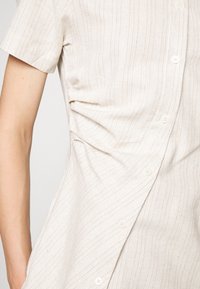 Camicia chiara con sottili righe, dotata di chiusura a bottoni e un design drappeggiato, mostrata parzialmente sulla parte superiore del corpo di un modello.
