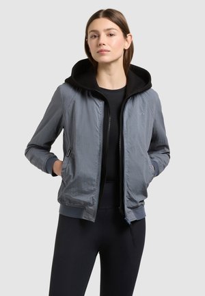 Jonge vrouw met een zwarte hoodie en een grijze jas met rits, met handen in de zakken, tegen een eenvoudige lichte achtergrond.