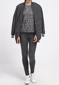Giubbotto bomber nero con polsini a strisce bianche, maglione in maglia grigia texturizzata, jeans skinny neri e sneakers bianche. Outfit casual a strati.
