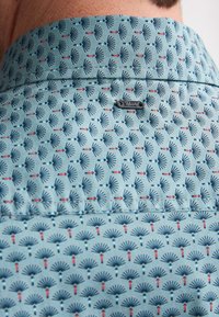 Camicia azzurro chiaro con un motivo a ventaglio ripetuto in blu navy e rosso. Include un piccolo logo vicino al colletto. Tessuto di texture liscia.