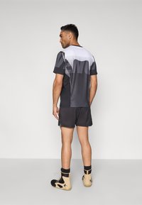 Atlétikai ruházat fekete és szürke rövid ujjú pólóval, amelyben átmenetes dizájn látható, fekete rövidnadrággal és bézs színű magas szárú cipőkkel párosítva.