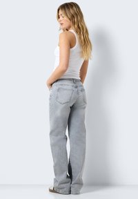 Mulher com cabelo loiro longo, vestindo uma camisola branca sem mangas e jeans retos cinza claro, de pé com as mãos nos bolsos, de lado.