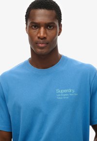 Blå bomulds t-shirt med korte ærmer, prydet med et diskret logo print i lys grøn på venstre bryst, samt en rund halsudskæring.