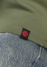 Camiseta de algodón verde oliva con un parche negro que presenta un acento circular rojo, colocada sobre una tela de denim azul texturizada.