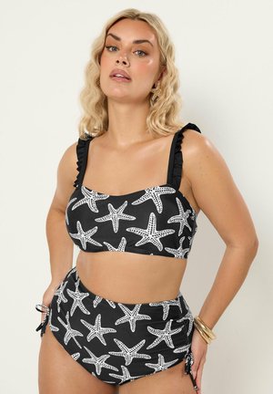 Donna bionda che indossa un bikini nero a vita alta con motivo a stella marina bianca, spalline arricciate, braccialetti dorati e orecchini a forma di stella.