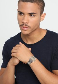 Montre numérique argentée avec un cadran octagonal, des accents noirs et un bracelet en métal. Portée au poignet d'une personne ayant les cheveux bruns courts et portant une chemise neutre.
