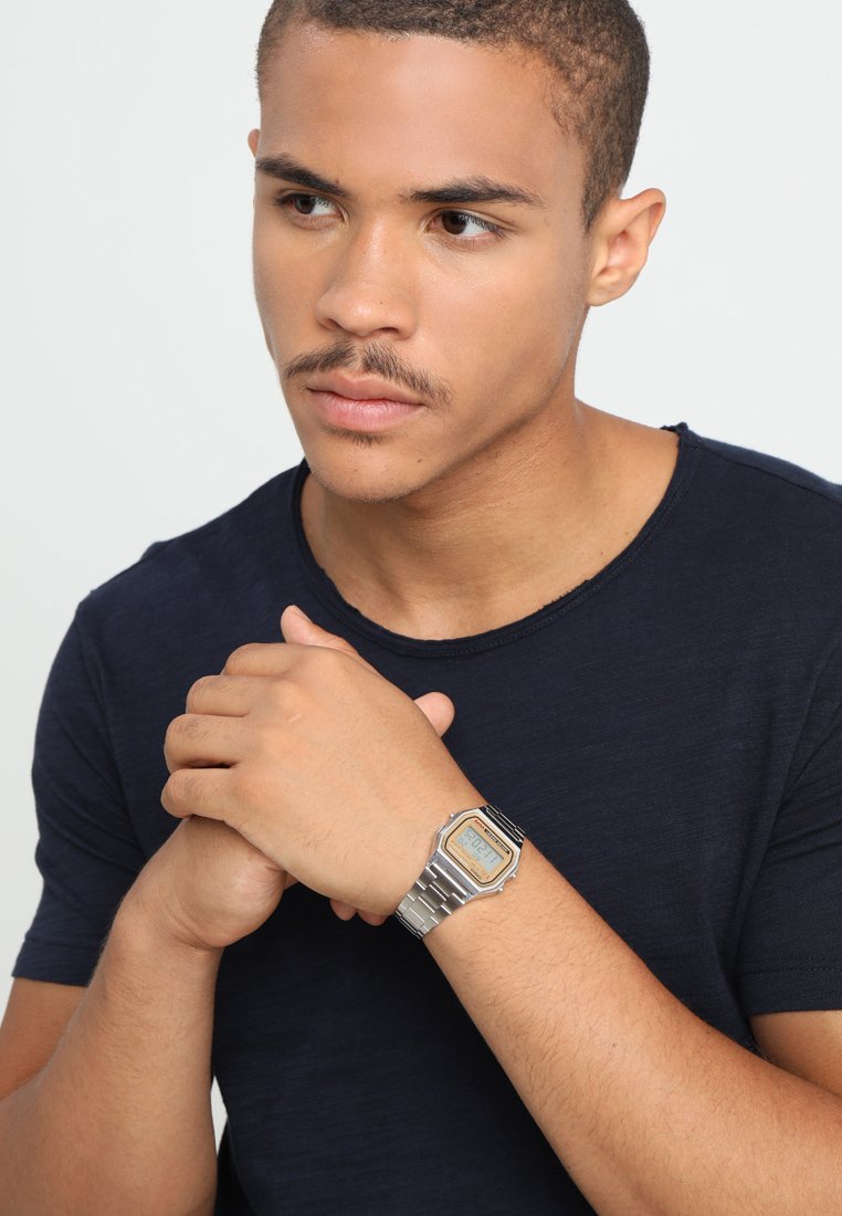 Montre numérique argentée avec un cadran octagonal, des accents noirs et un bracelet en métal. Portée au poignet d'une personne ayant les cheveux bruns courts et portant une chemise neutre.