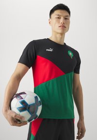 Camiseta de fútbol de manga corta en negro, rojo y verde con bloques de color en ángulo; presenta el logo de Puma; el jugador sostiene un balón de fútbol con un patrón.