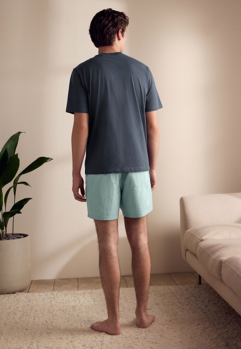 Tenue décontractée pour homme comprenant un T-shirt gris foncé et un short vert clair texturé. Intérieur avec une plante et des coussins doux.