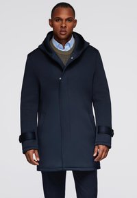 Abrigo navy con un alto cuello, tejido suave, cierre frontal a presión, dos bolsillos laterales y puños ajustables. Llevado sobre un suéter y una camisa.