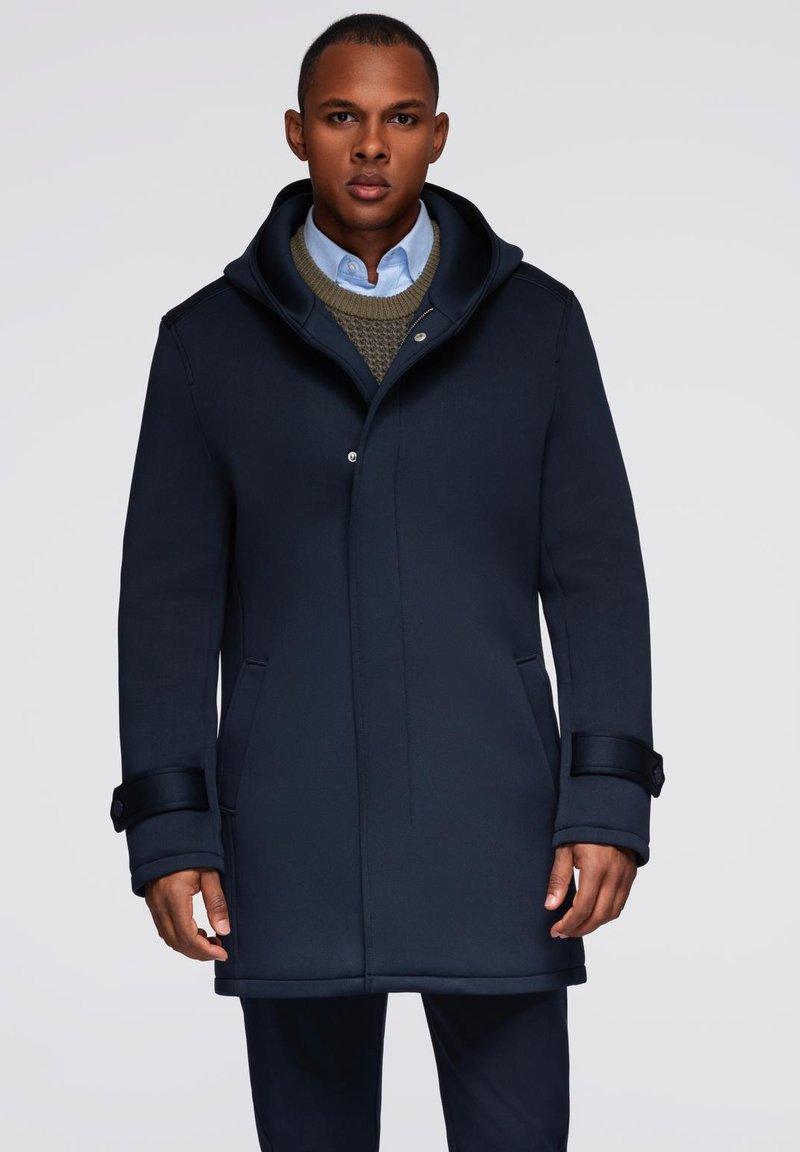 Abrigo navy con un alto cuello, tejido suave, cierre frontal a presión, dos bolsillos laterales y puños ajustables. Llevado sobre un suéter y una camisa.