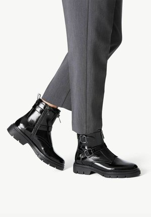 Black patent leather ankle boots with zippers, buckle accents, and textured soles, paired with gray tailored trousers.