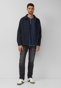 Dunkelblaues Button-up-Jacket aus strukturiertem Stoff, über einem marineblauen Henley-Shirt getragen, kombiniert mit grauen Jeans und weißen Sneakern mit Akzenten.