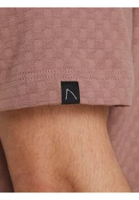 Krótki rękaw z fakturowanej, mauve tkaniny z małą czarną metką z prostym, białym, haftowanym wzorem geometrycznym.