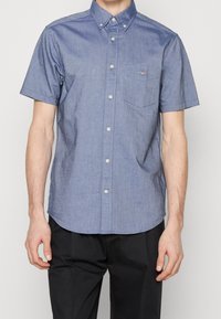 Homme portant une chemise chambray bleue à manches courtes avec une poche et un pantalon noir sur un fond uni.