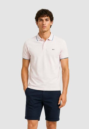 Hombre con camiseta polo rosa claro con ribete azul y pantalones cortos azul marino, de pie con una mano en el bolsillo frente a un fondo liso.