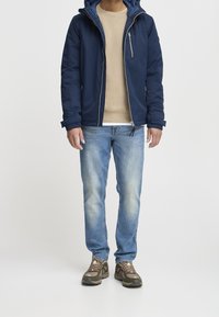 Giacca blu navy con cappuccio e zip frontale, abbinata a un top beige, jeans azzurri chiari e scarpe da trekking marroni.
