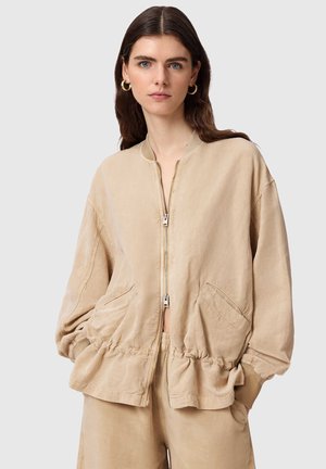 Ung kvinde med langt brunt hår, der bærer gyldne øreringe og en beige lynlåstrøje med indsnøret talje og matchende bukser.