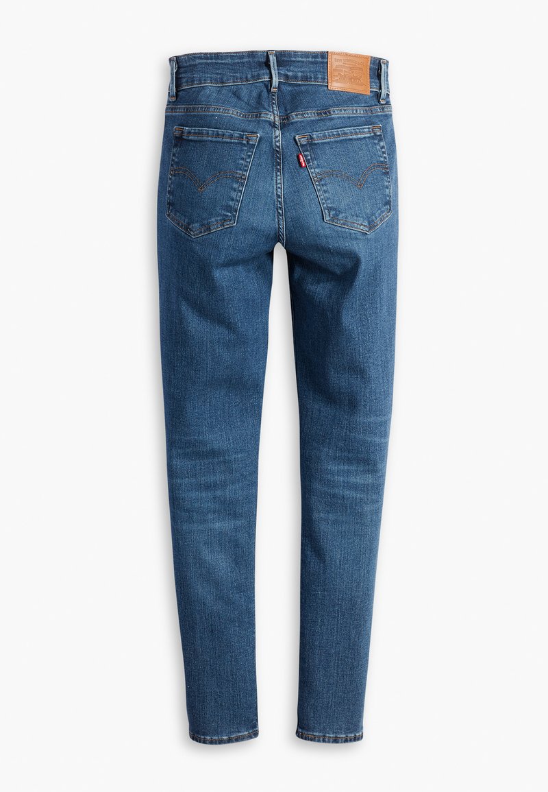 Jean en denim bleu avec une coupe ajustée, cinq poches, coutures visibles et une étiquette en cuir marron à la taille arrière.