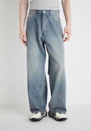 Person wearing loose-fitting faded blue jeans and white sneakers with black soles against a plain background.