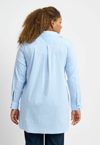 Light blue long-sleeve shirt with a collared neckline, featuring a pleat at the back and buttoned cuffs. Made from smooth fabric.