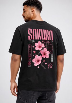 Man wearing a black T-shirt with large pink cherry blossom design and text "SAKURA" and Japanese characters on the back.