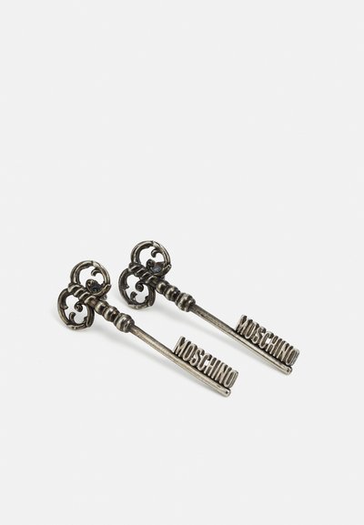 Duos išskirtinių metalinių raktų, puošnių su sudėtingais raižiniais rankenomis ir su užrašu „MOSCHINO“ ant ilgo stiebo, eksponuojami ant baltos paviršiaus.